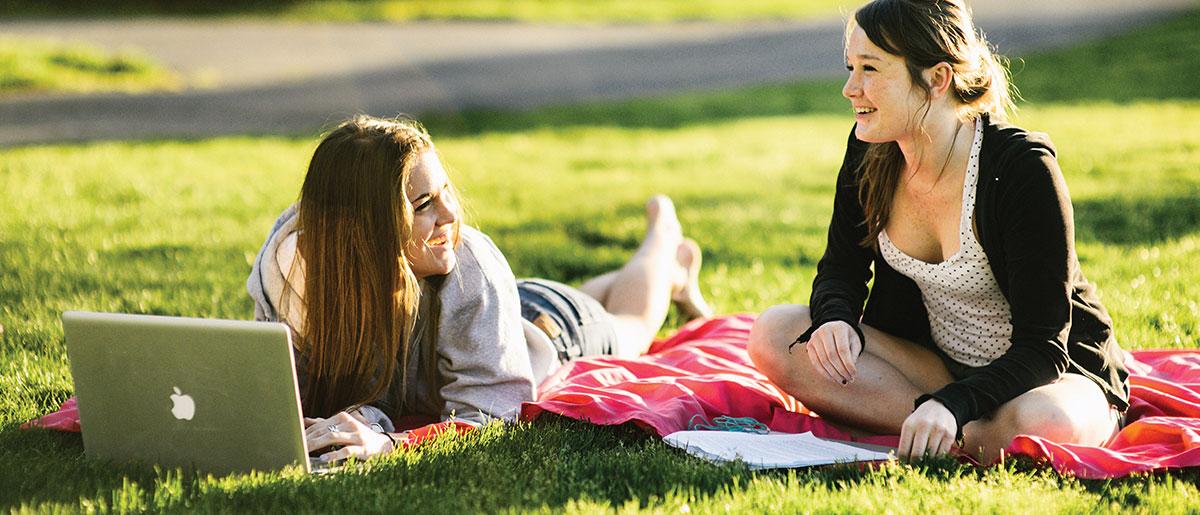 Students relaxing on the grass, on is laying on a blanket in front of a computer.